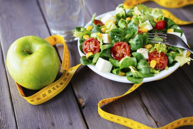 Fitness salad and apple fruit surrounded by measuring tape on rustic wooden table. Mixed greens, tomatos, diet cheese, olive oil and spices for healthy lifestyle concept.